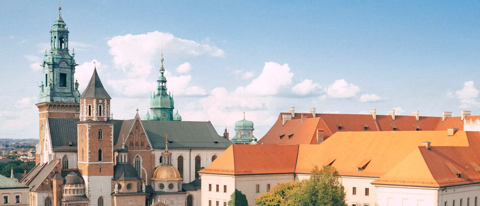 Wawel Cathedral seen from above — the copper-green Clock Tower and cupolas, the gilded dome of the Sigismund Chapel and the long red-tiled roofs of the castle ranges under a cloud-flecked sky