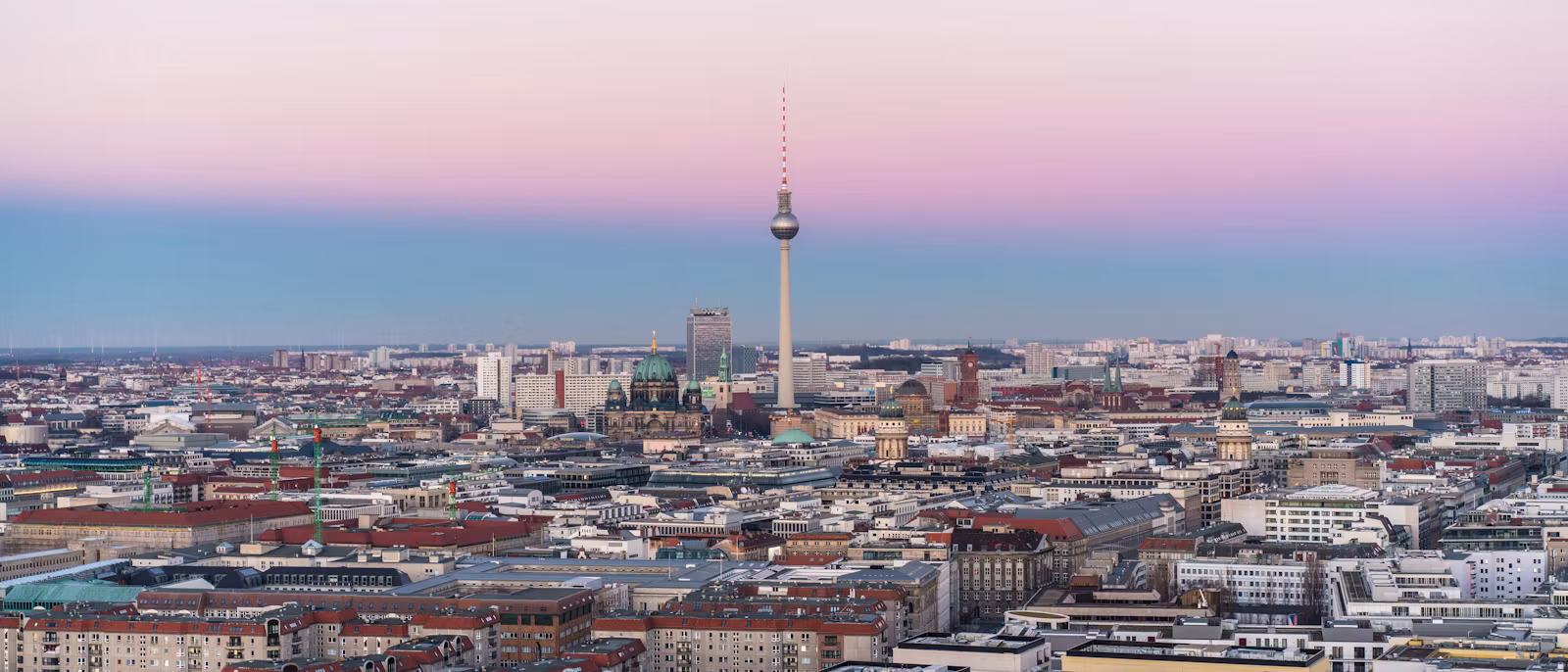 Berlin at dusk from a high viewpoint — the Fernsehturm rising out of a sea of roofs under a pink and blue sky