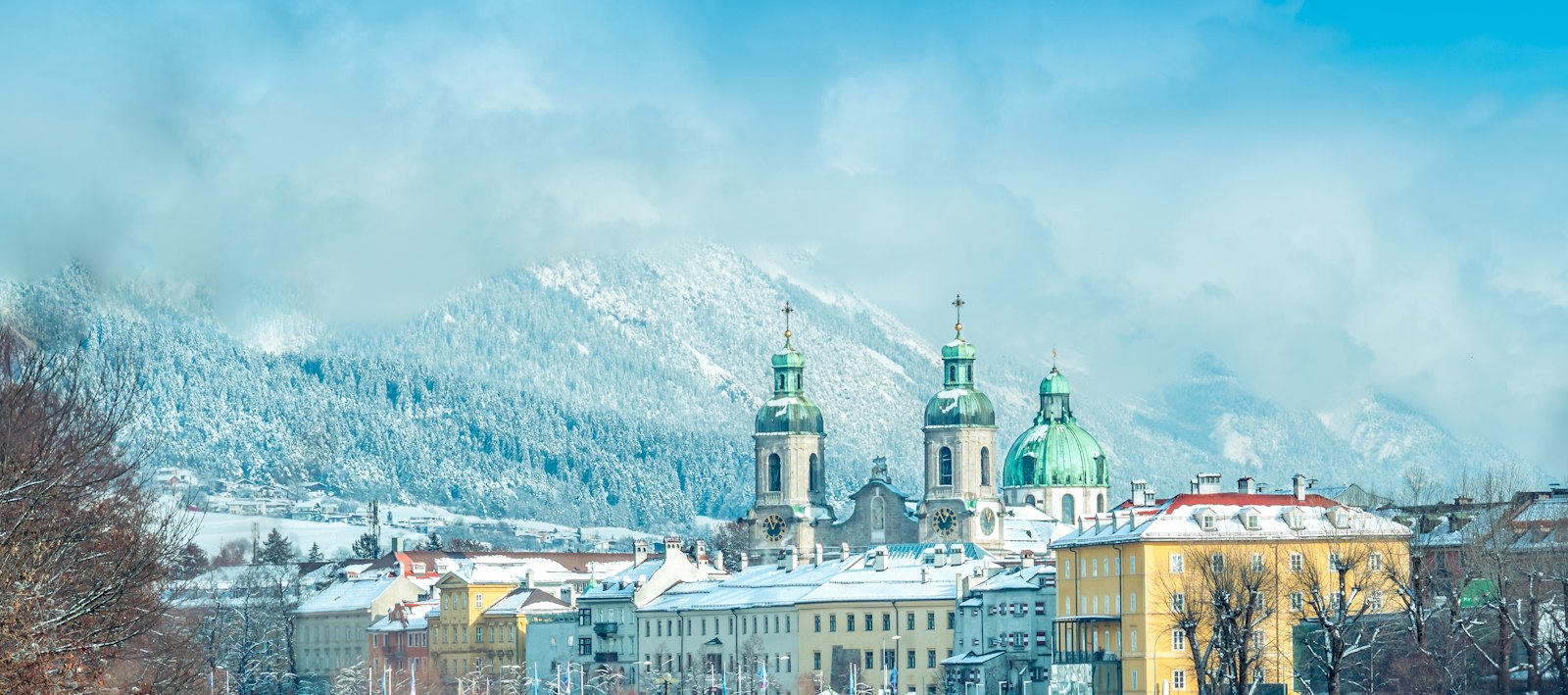 Snow-covered Innsbruck rooftops and the twin towers and green dome of the cathedral, with the forested, snow-dusted Nordkette rising behind the city
