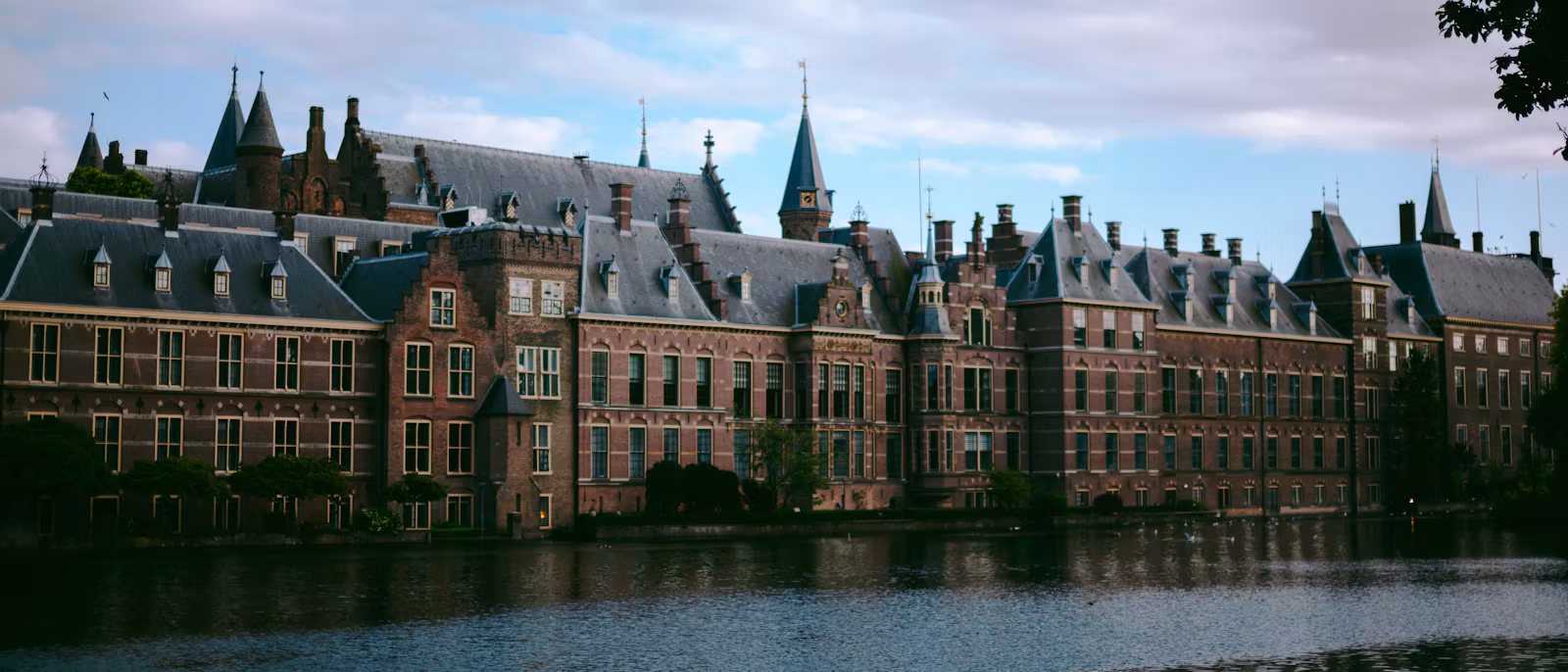 The Binnenhof seen across the Hofvijver — a long row of red-brick façades, slate roofs, turrets and spires reflected in the dark water under a sky of white clouds