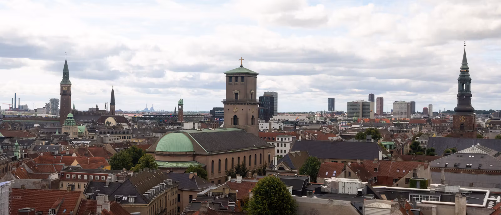 Copenhagen from above — the green copper dome and square tower of the Church of Our Lady, the City Hall clock tower at the left and the spire of St Nikolaj at the right, over red and slate roofs under a cloudy sky